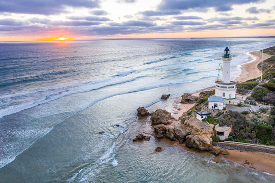 Aerial view of the sun setting off shore from a lighthouse on a cliff top