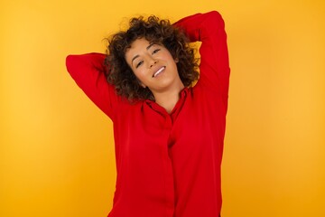 Young arab woman with curly hair wearing red shirt  on yellow background stretching arms, relaxed position.