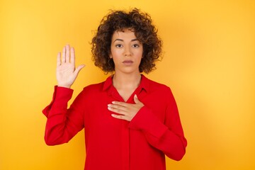 Young arab woman with curly hair wearing red shirt  on yellow background Swearing with hand on...