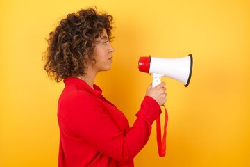 Young arab woman with curly hair wearing red shirt holding a megaphone over yellow background looking to side, relax profile pose with natural face with confident smile.