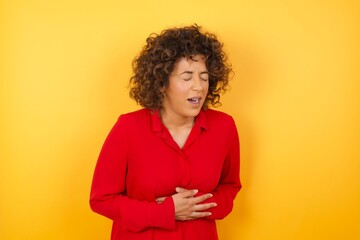 handsome young man wearing red shirt over yellow isolated background with hand on stomach because nausea, painful disease feeling unwell. Ache concept.