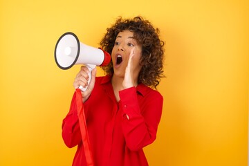 Young arab woman with curly hair wearing red shirt holding a megaphone over yellow background shouting and screaming loud to side with hand on mouth. Communication concept.