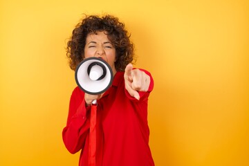 Young arab woman with curly hair wearing red shirt holding a megaphone over yellow background...