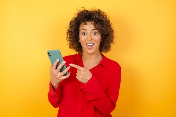 Portrait of  arabn businesswoman holding in hands showing new cell, wearing red shirt standing against yellow background