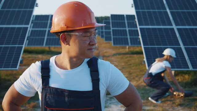 Portrait of cheerful adult asian enginner in industry uniform cooperating on ecological construction of solar farm outdoor. Workman. Renewable energy. Solar field.