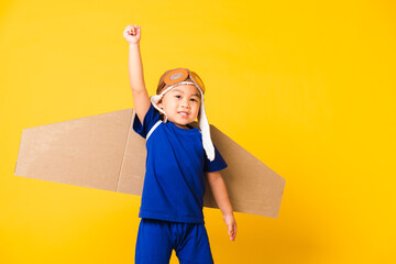 Happy Asian handsome funny child or kid little boy smile wear pilot hat play and goggles raise hand up with toy cardboard airplane wings flying, studio shot isolated yellow background, Startup freedom