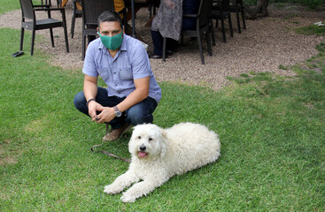 Latino man with protection mask walking on the grass with white dog at the lake shore, new normal