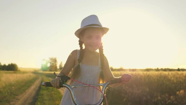 Happy child rides a bicycle in the park. A girl in a dress pedals in the sun. The kid is enjoying the dream. Cheerful child expresses emotions of happiness.