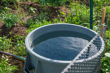 A blue sky, puffy clouds and a wire fence are reflected in a large barrel of irrigation water in a fenced in garden.