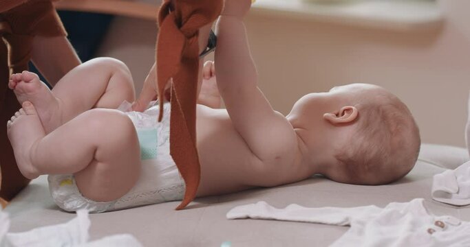 Close up of an adrorable baby, lying on its back on the couch, holding a pacifier, adn its' mother's hands, who changes the diapers for baby