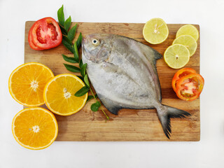 top view of Black Pomfret Fish dish cooking with various ingredients. Fresh raw fish decorated with lemon slices and tomato slice and curry leaves on a wooden pad,white background.