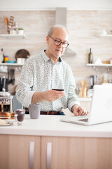 Elderly man holding credit card, using modern payment system. Pensioner paying online using credit card and application from laptop during breakfast in kitchen. Retired elderly person using internet