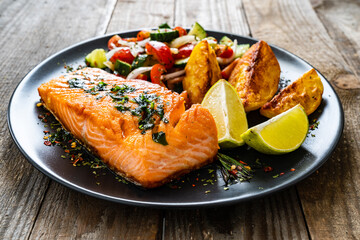 Fried salmon fillet with fried potatoes, lime and vegetable salad served on black plate on wooden table
