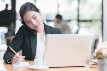 Portrait of young beautiful businesswoman using smartphone while sitting and working with laptop in contemporary office room.