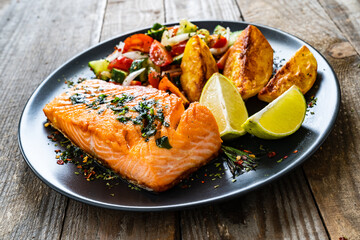 Fried salmon fillet with fried potatoes, lime and vegetable salad served on black plate on wooden table
