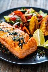 Fried salmon fillet with fried potatoes, lemon and vegetable salad served on black plate on wooden table
