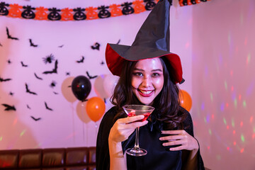 Portrait of young Asian woman smiling in Halloween costumer as witch in party, holding wine glass, looking at camera.