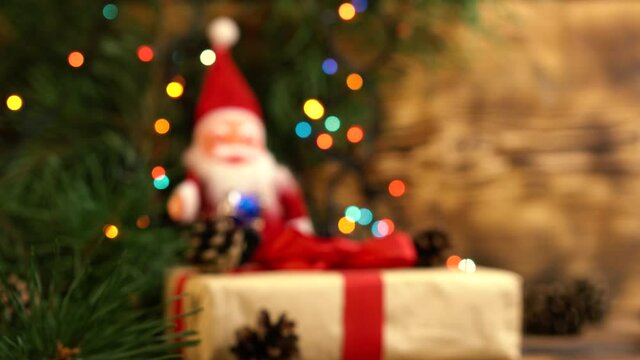 plush red santa claus with a big white beard, sits on a gift under fir branches and a multicolored gerland, blurred image, christmas blurred background. Close-up