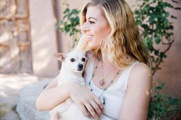 Young blond haired woman holding cuddling a blond short haired Chihuahua dog in her arms with loving affection.  She is outdoors in a southwestern beige and light green environment.  She is wearing an