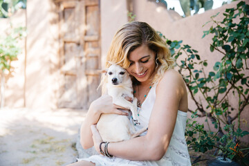 Young blond haired woman holding cuddling a blond short haired Chihuahua dog in her arms with loving affection.  She is outdoors in a southwestern beige and light green environment.  She is wearing an