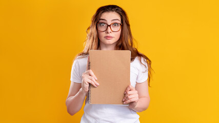 Scared student portrait. Exam anxiety. Worried woman in glasses with beige blank notebook isolated on orange copy space background. Panic attack. Higher education.