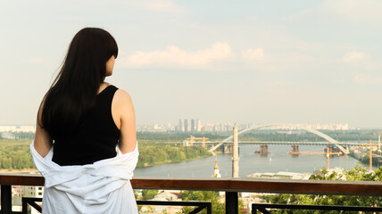 Back view of a young woman in a white shirt, a brunette traveler looks at the cityscape on a sunny day from a high hill with an observation deck for tourists. Vintage