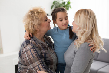 Naklejka premium Three generations of women. Beautiful woman and teenage girl are kissing their granny while sitting on couch at home