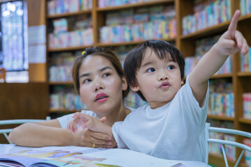 Asian young mother with little boy reading book together in library. Happy family. Parent educating children. Pre-school learning concept.