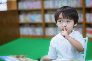 Asian little boy reading book and holding his finger on lips making a silent gesture in library. Pre-school learning concept.
