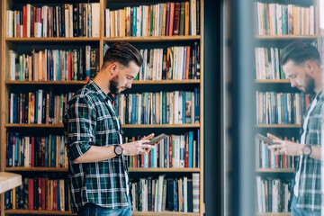 Stylish male student standing with book near bookcase in university library