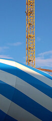 Close-up of the back of a blue white striped cement mixer truck in front of a yellow crane and the...