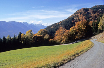 Fototapeta na wymiar Single track roadway winding down from mountain summit towards valley. Autumn leaves on forest trees on hillsides. Carnic mountain range on horizon. Blue sky and clouds