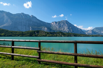 Fototapeta na wymiar Cavedine Lake . Panorama of the turquoise blue waters, with a wonderful wooden fence, rich vegetation and mountains. Cavedine Lake is a small alpine lake in Dolomiti Trentine, Italy.