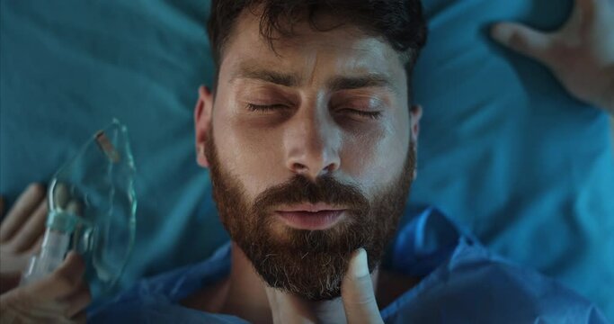 Close up of medics hands putting on ill bearded man oxygen inhalation mask. Top view of unconscious guy in 30s mumbling while lying on medical transportation cart on way to hospital