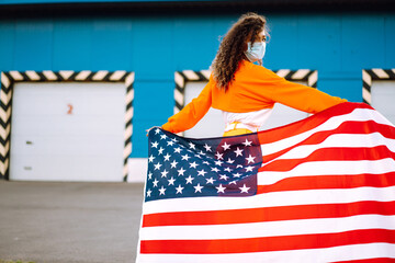 African American woman outdoors, wearing a protective mask against coronavirus and holding a USA flag.