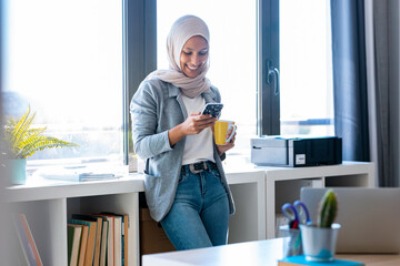 Beautiful young muslim business woman wearing hijab using her smart phone while standing next to the window in the office.