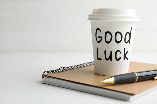 Paper coffee cup with phrase GOOD LUCK and stationery on white wooden table, closeup. Space for text