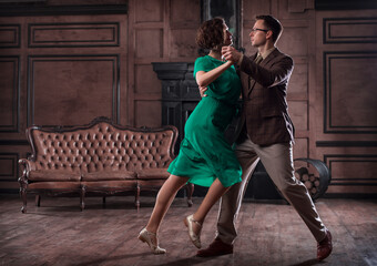 a young couple dancing in a retro hall in front of a fireplace and a leathern sofa; the woman wears a green dress
