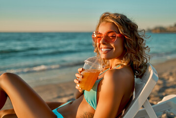 Attractive caucasian woman in sunglasses and a swimsuit drinks juice or lemonade and sunbathes while lying on a sun lounger on the beach.