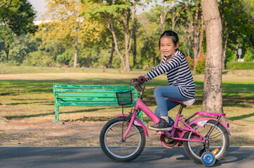 Activity morning in public park Thailand, Asian child girl practicing biking bicycle with two supporter wheels, first time with bicycle. Smiling face child on bicycle.