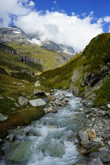 Paesaggio di Montagna, si vede l'acqua del fiume scorrere in mezzo alla natura tra le roccia e l'erba verde, sopra il cielo azzurro con le nuvole bianche e sullo sfondo il ghiacciaio alpino 