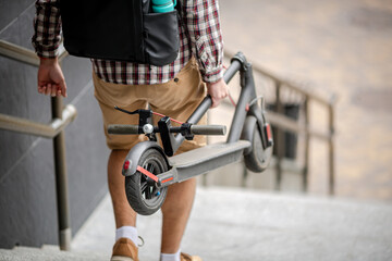 Young caucasian man in casual clothes with a backpack carries a folded electric scooter in his hand...