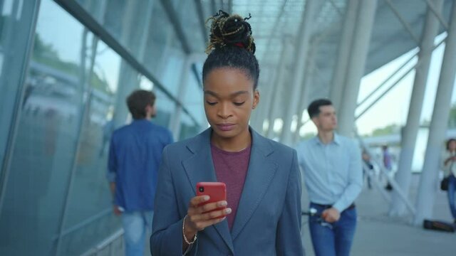 Business casual afro-american female manager holding smartphone communicating in social media walking outdoors. Airport building. Business center. Crowd.
