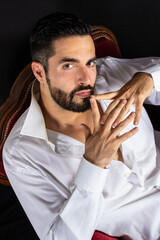 Portrait of handsome seductive man with open shirt showing chest sitting on a luxury red armchair. Attractive stylish bearded man in formal wear. Black background. High angle.