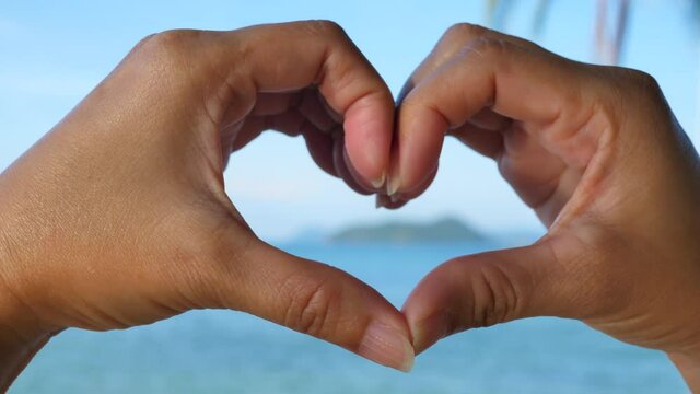 Love shape hands at the beach. Female hand making a heart shape against a beautiful blue sky.