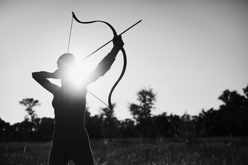 Young Caucasian female archer shooting with a bow in a field at sunset.