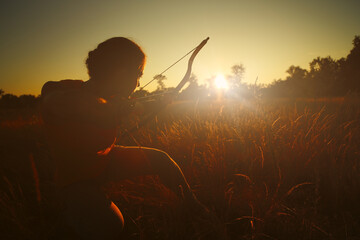 Young Caucasian female archer shooting with a bow in a field at sunset.
