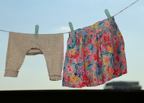 Baby girl clothes or tiny grey pants and colorful flower motif dress drying on a wire with blurred city building in the background