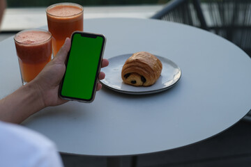 over the shoulder view of people holding green screen mobile phone. With bread and fruit juice on white table.