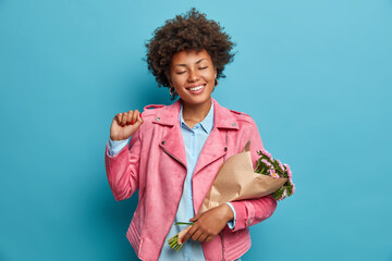 People spring holiday and harmony concept. Pretty smiling woman with Afro hair raises clenched fist, enjoys good time of life. Positive lady in fashionable pink jacket holds bouquet of flowers